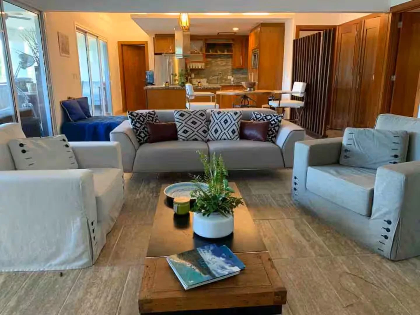 Sala de estar con sofá gris, sillones blancos, mesa de madera con plantas y libros, fondo cocina con barra y sillas, ventanas grandes con vista exterior y piso de baldosas.