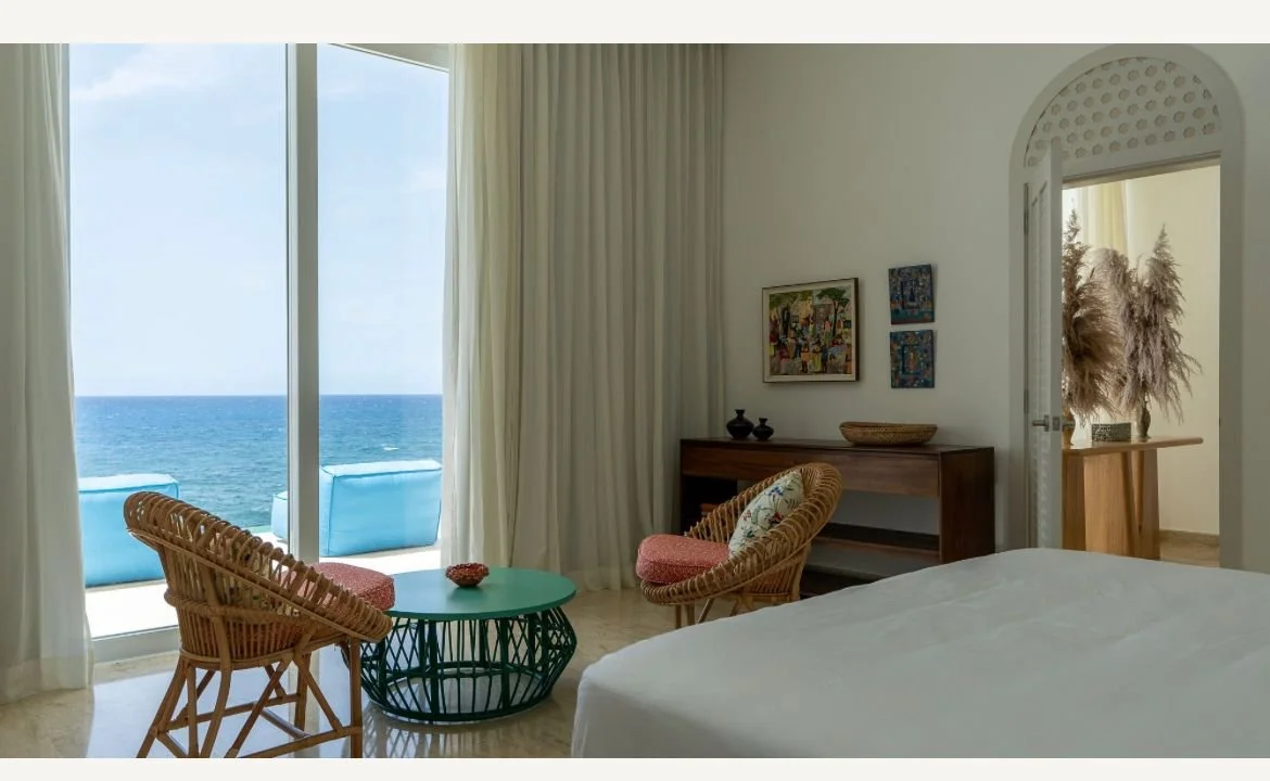 Habitación luminosa con vista al mar, muebles de mimbre, mesa verde, obras de arte en la pared y puerta con jarrón de pampas.