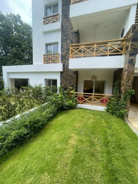 Exterior view of a modern apartment building with multiple balconies and a small green lawn. The balconies feature wooden railings, and the facade includes a mix of white and stone elements. Lush greenery surrounds the building, creating a tranquil o