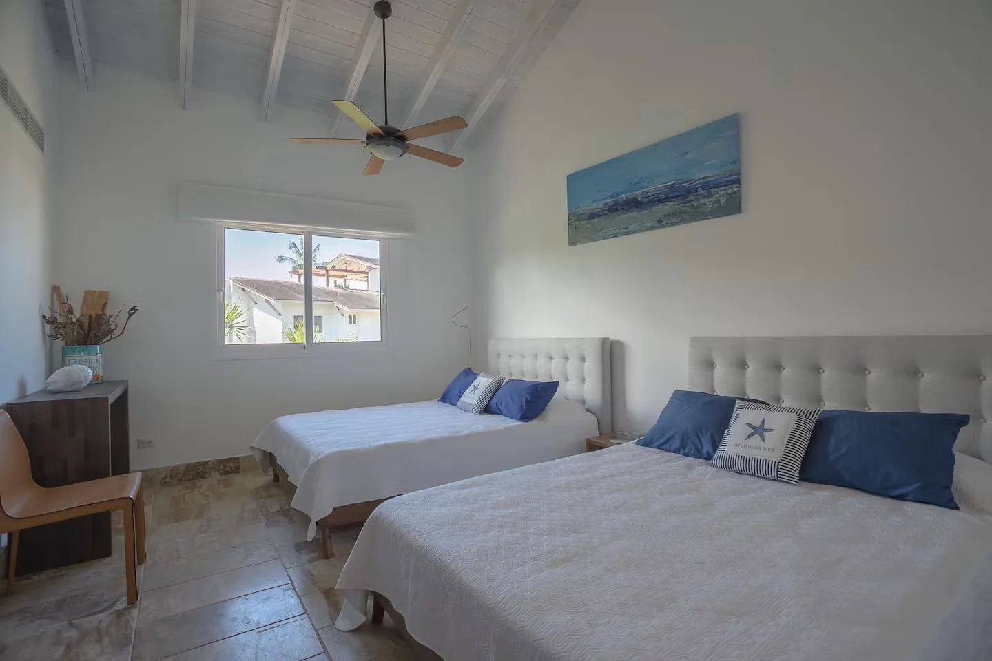 Habitación con dos camas con cabecera blanca, almohadas azules y decorativas con tema de playa, pintura de paisaje marítimo en la pared, ventana con vista a casas y palmeras, ventilador en el techo