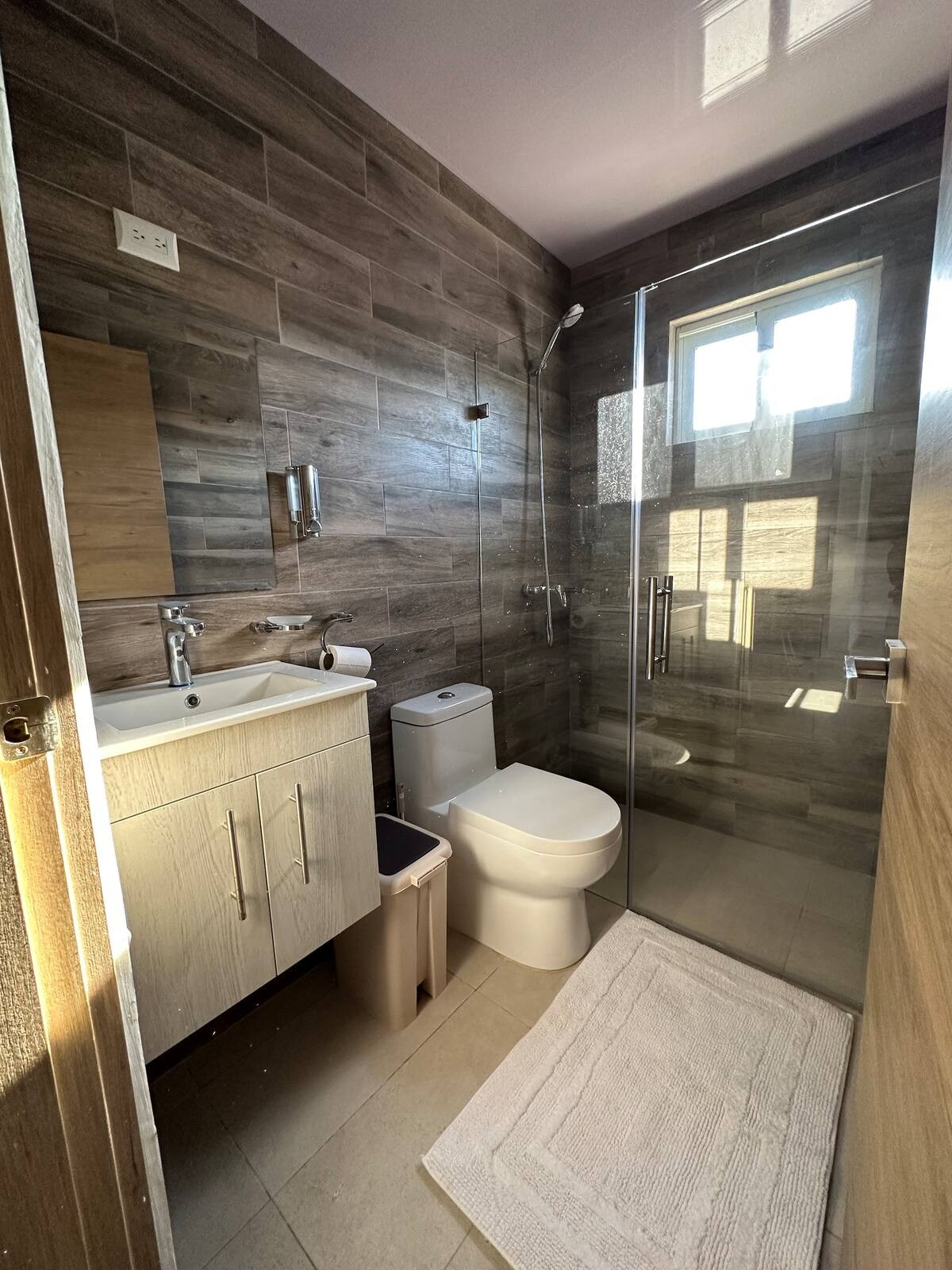 Modern bathroom with a wood-paneled design, featuring a white vanity with a sink and cabinet, a toilet, a glass-enclosed shower, and a beige floor mat.