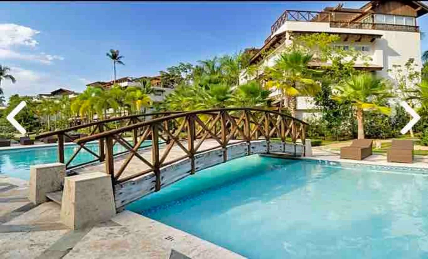 Piscina con puente de madera, rodeada de plantas y árboles, con un edificio en el fondo y cielo despejado.