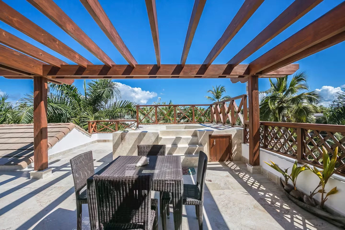 Terraza exterior con comedor al aire libre, muebles de mimbre y plantas tropicales, con cielo azul y nubes.