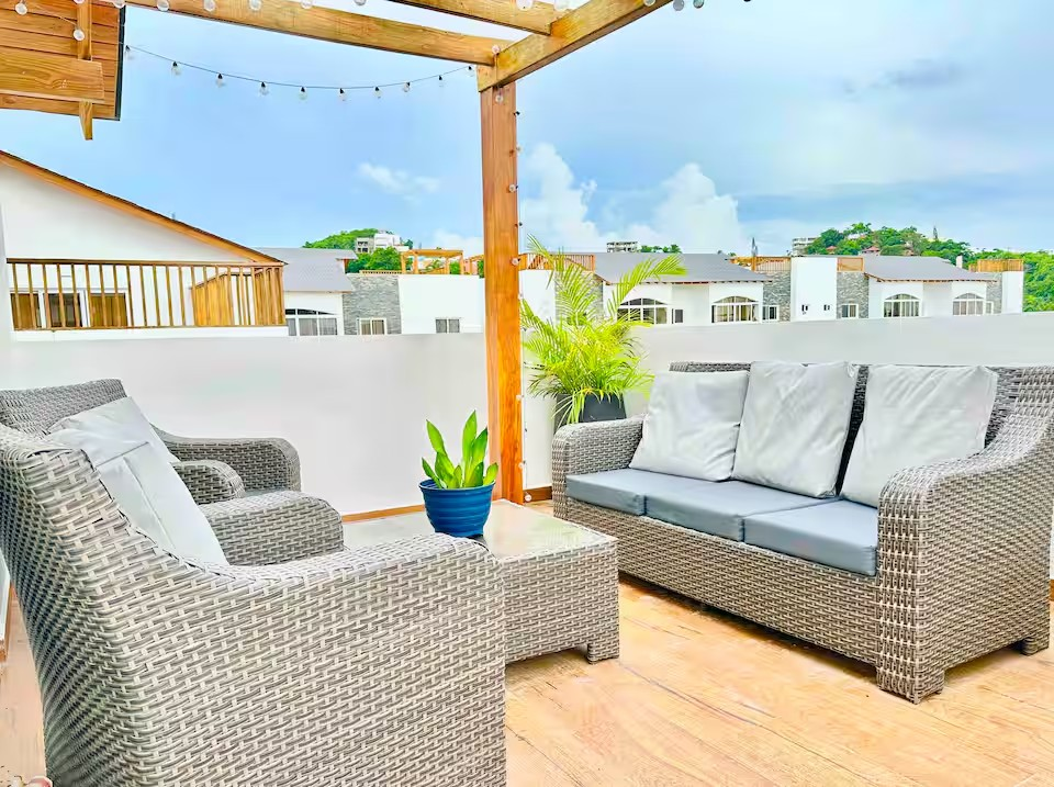 Outdoor patio with wicker furniture, including a sofa and chairs, under a wooden pergola with string lights, and potted plants, against a backdrop of residential buildings and a partly cloudy sky.