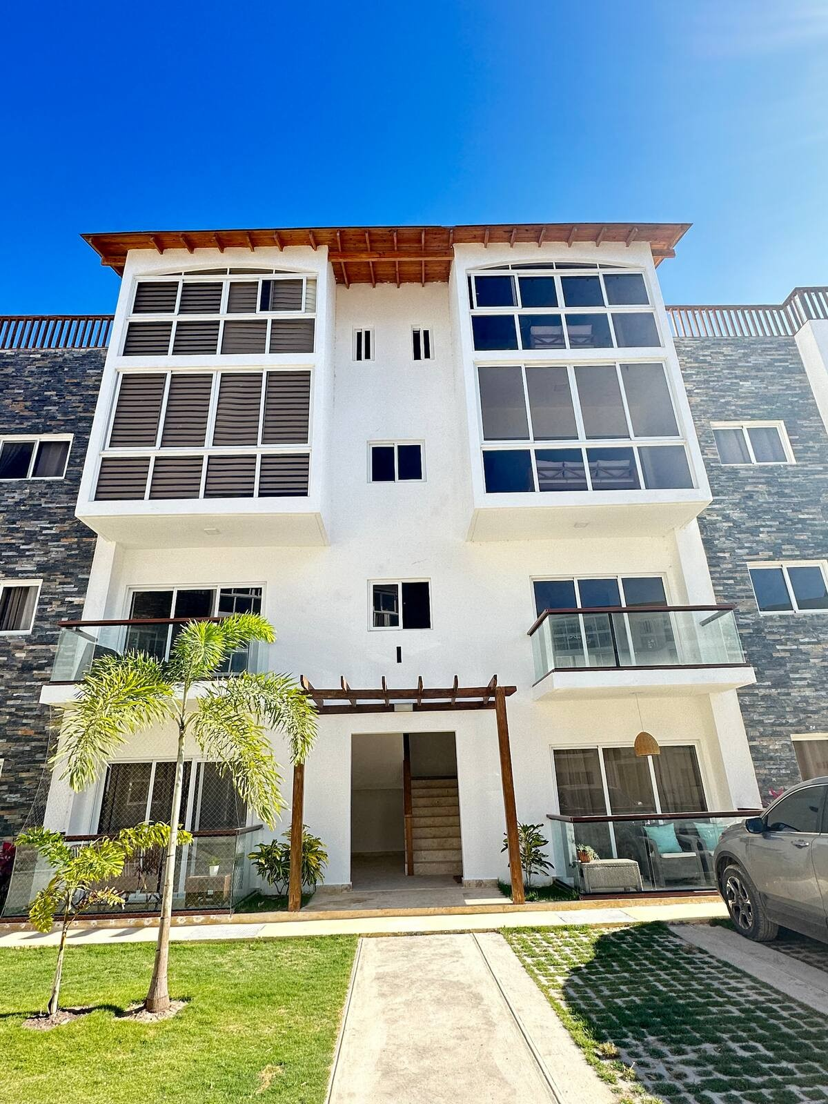 Modern three-story apartment building with stone facade, large windows, balconies, and a well-maintained lawn. Palm trees and a parked car are visible. Clear blue sky overhead.