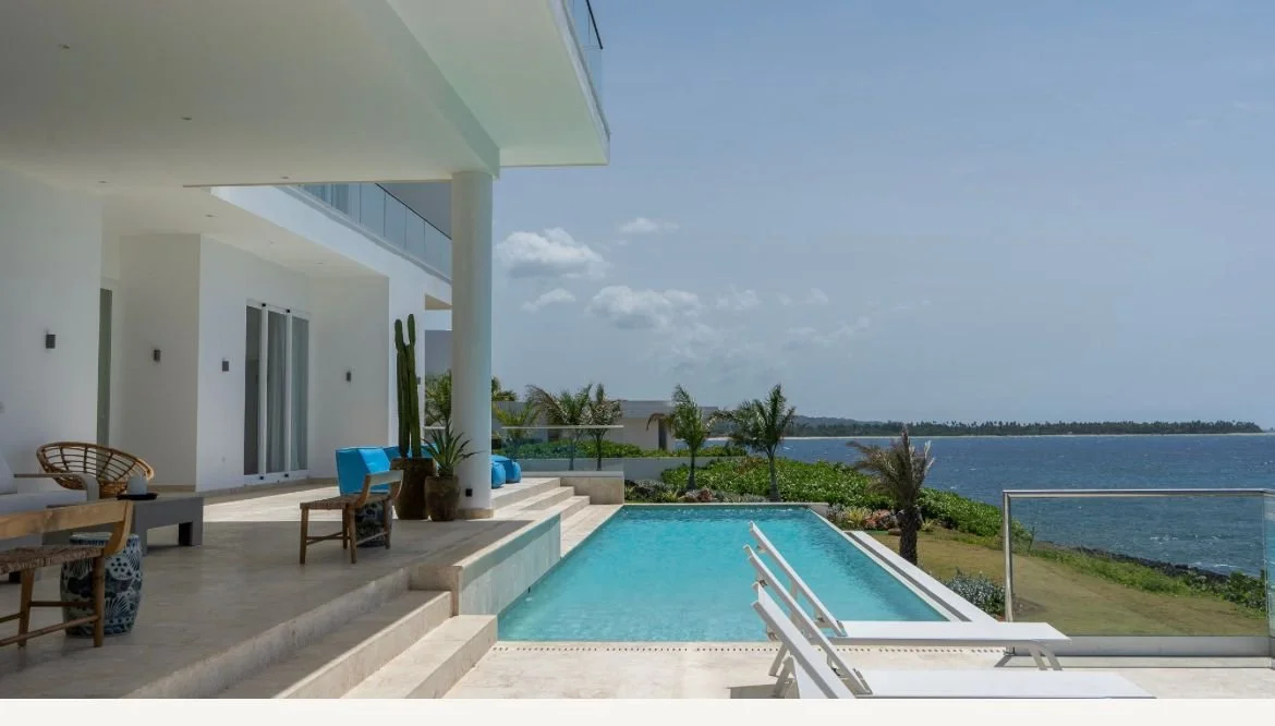Casa moderna junto a una piscina con vista al mar, rodeada de vegetación y cielo despejado, con sillas y mesas en la terraza.