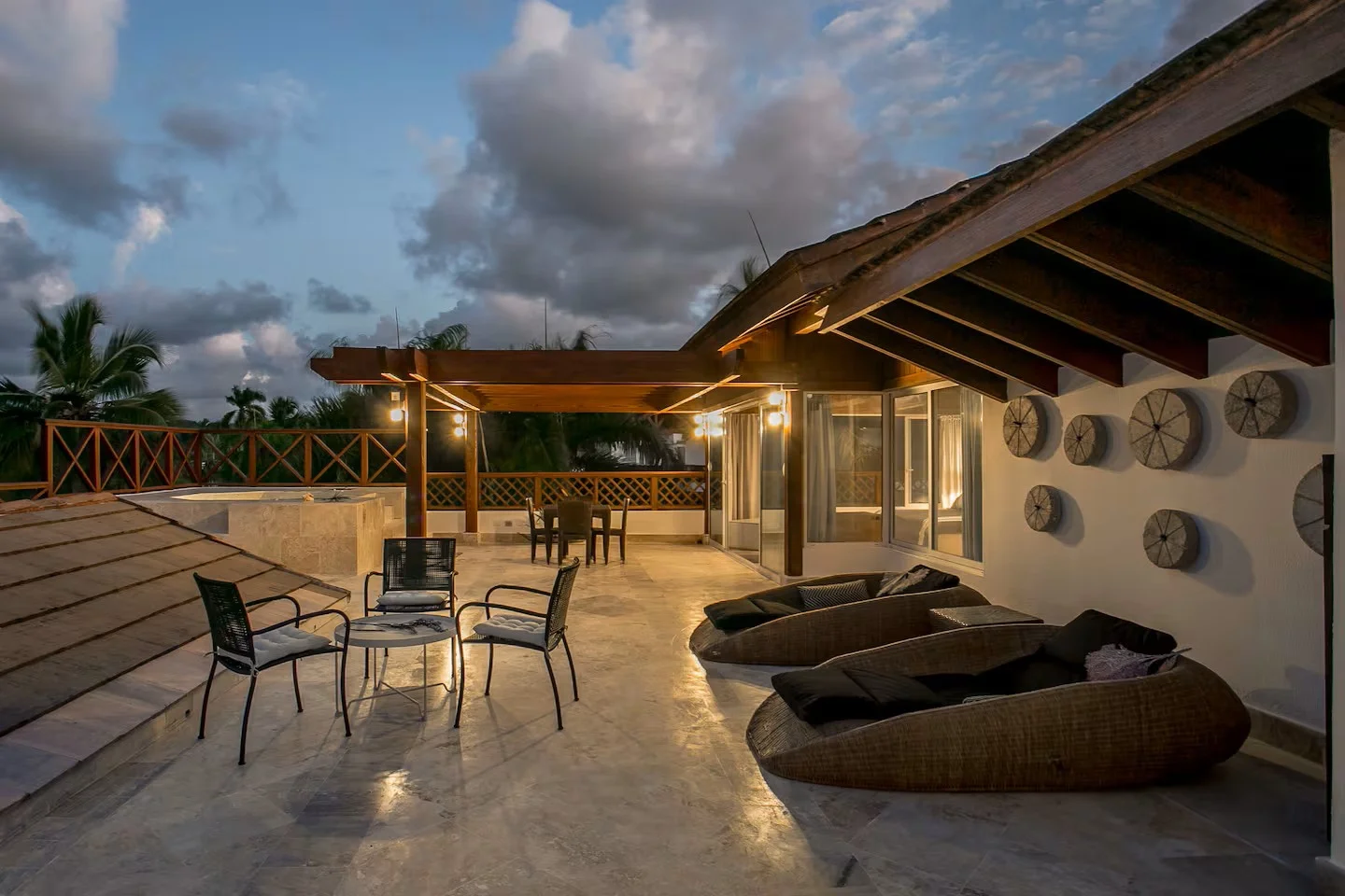 Terraza exterior con muebles de patio, sillas, mesas, techo de madera, paredes decoradas, vistas a árboles y cielo con nubes, ambientación nocturna.
