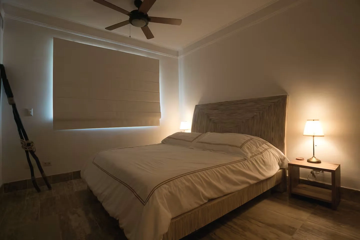 Habitación con cama grande, cabecera de madera, lámparas de mesita de noche, ventana con cortina cerrada, ventilador en el techo y suelo de madera.