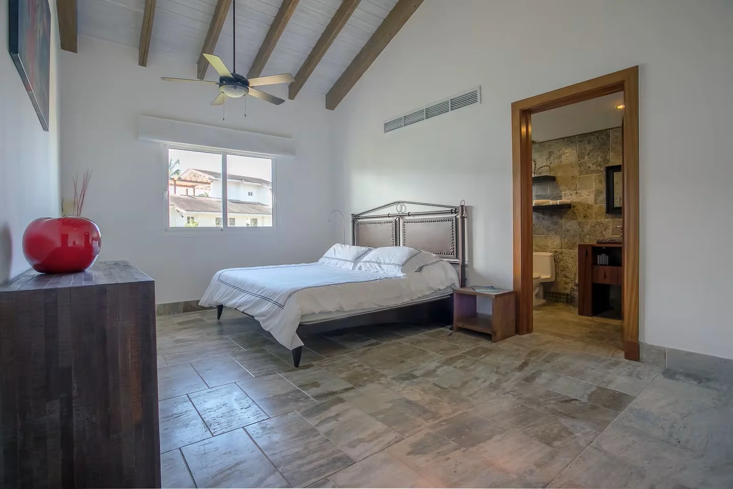 Habitación con cama, ventana y baño adjunto, decorada con madera y tonos neutros, piso de baldosas y techo de madera con ventilador.