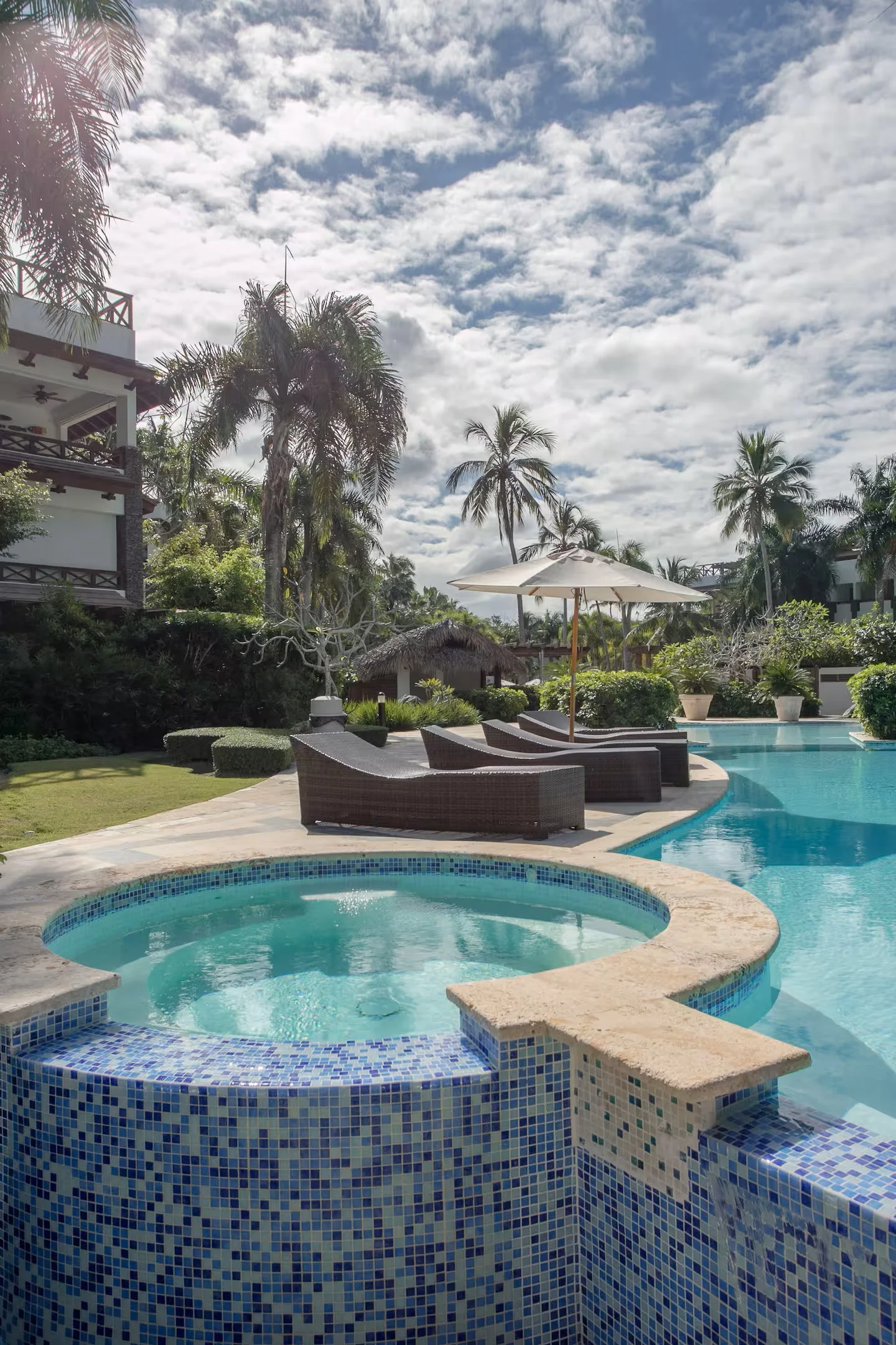 Piscina y jacuzzi en un entorno tropical con palmeras, sombra de paraguas y áreas de descanso, en un día soleado con cielo parcialmente nublado.