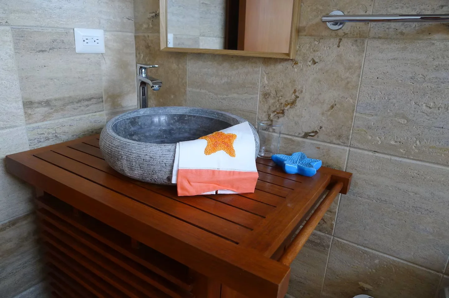 Lavabo de piedra en encimera de madera en un baño con pared de azulejos beige. Un paño decorativo en forma de estrella sobre el lavabo, una taza de plástico azul y una taza de vidrio en la encimera.