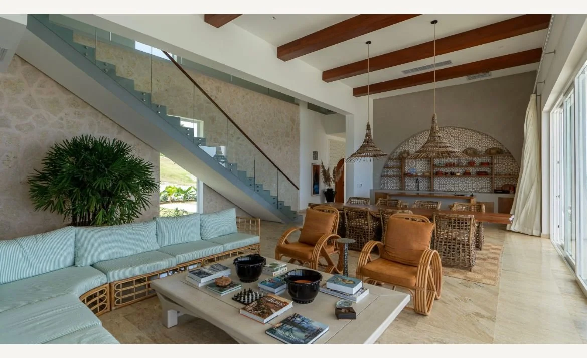 Sala de estar moderna con decoración rústica, sofá en forma de L, mesa de centro con libros y tablero de ajedrez, sillas de mimbre, lámparas colgantes de mimbre, escaleras de cristal y comedor en el fondo.