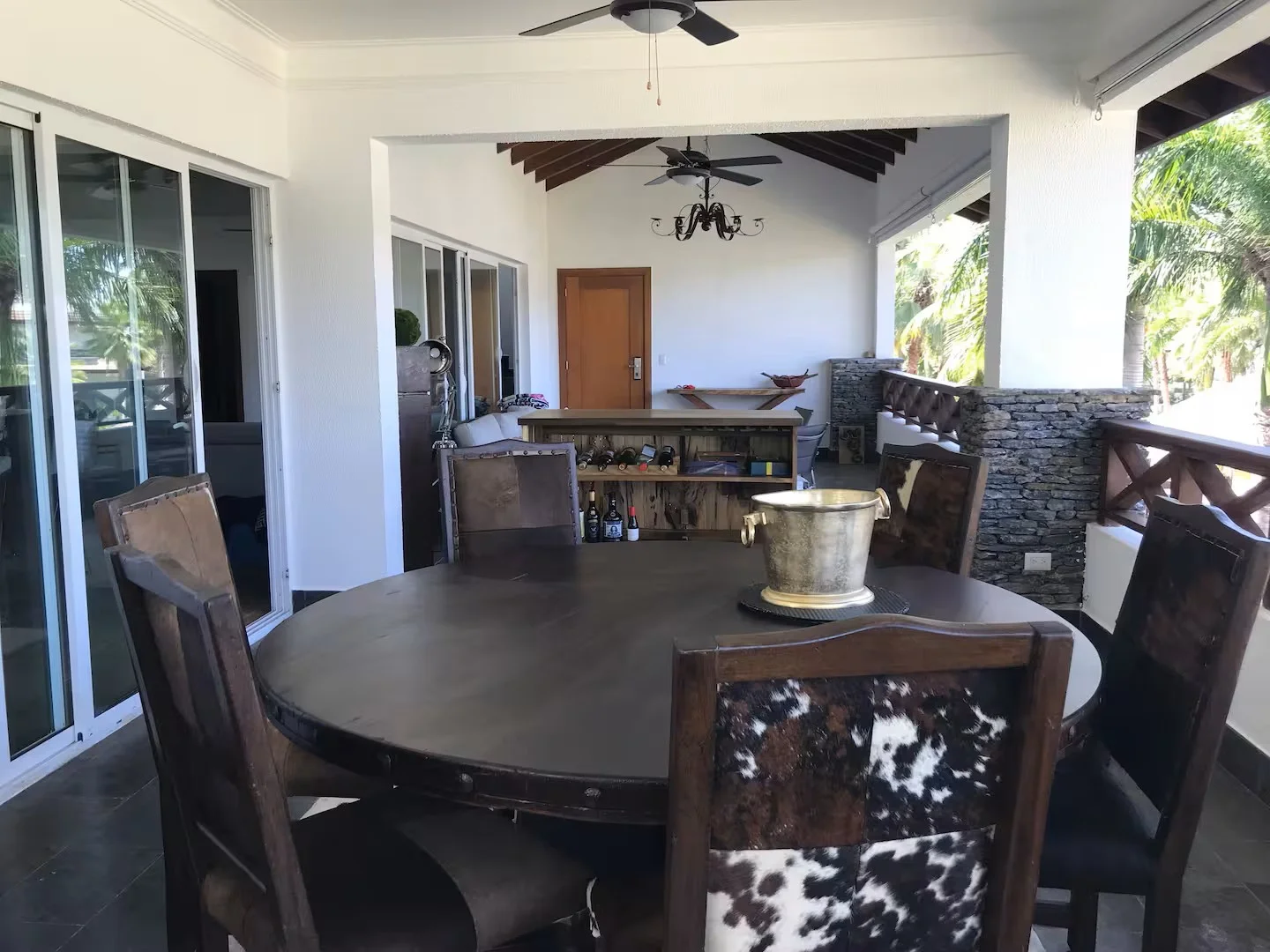 Área de comedor en un espacio abierto con mesa redonda de madera y sillas con asientos de cuero y caprino. Detrás, una barra con botellas y una decoración rústica, incluyendo una pared de piedra y ventiladores en el techo, rodeada de árboles verdes p