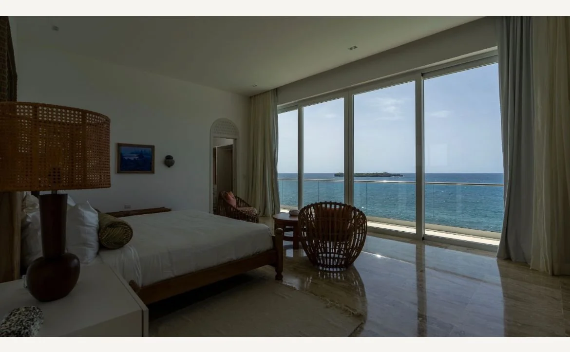 Habitación de hotel moderna con vista al mar, cama grande, lámpara y sillas de mimbre.