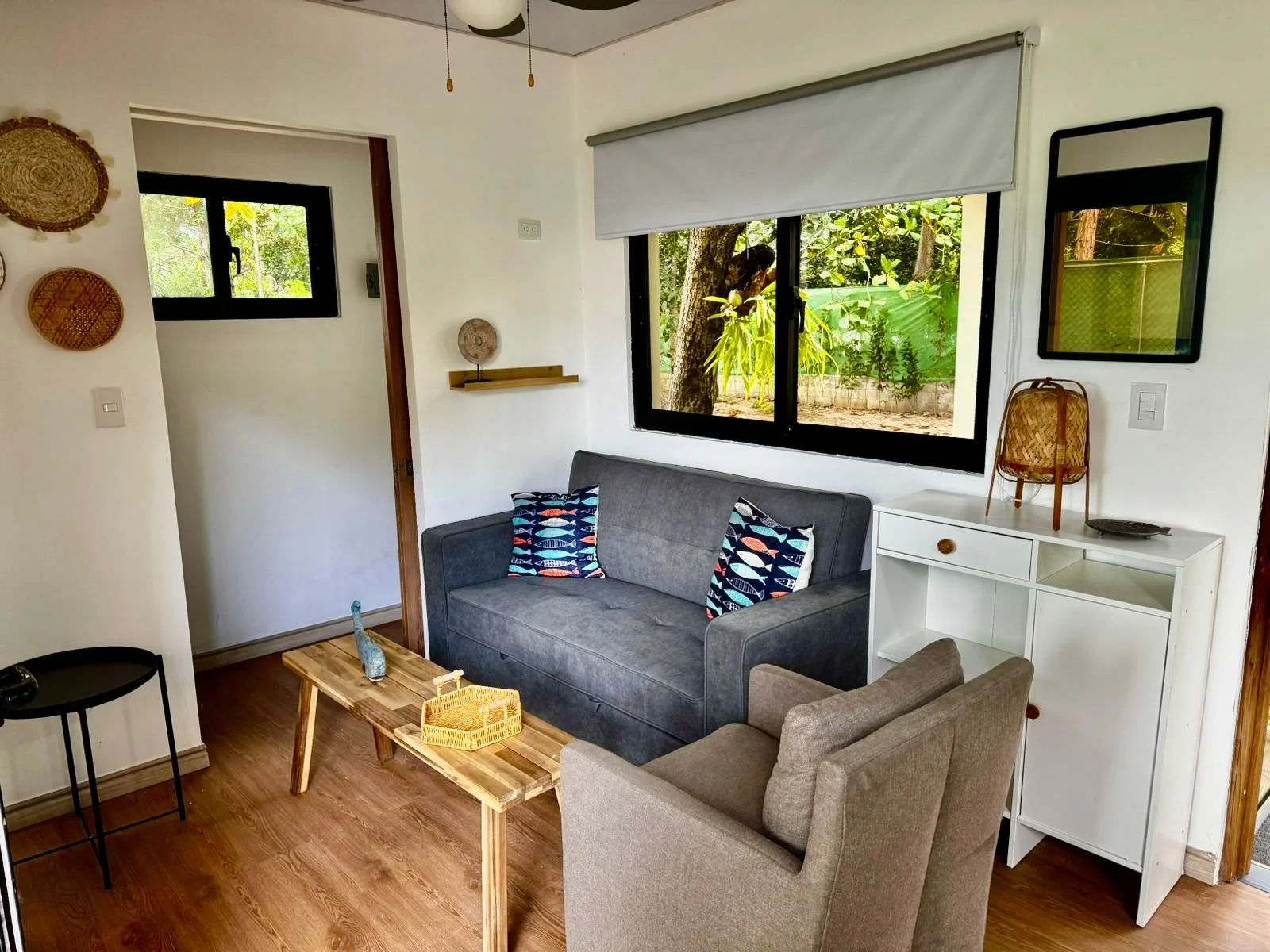 Sala de estar con sillón gris y sillón beige, mesa de madera, sofá con cojines con diseño de peces, ventana con vista a un jardín con árboles y plantas, decoración con cestas de mimbre en la pared y en la mesa, espejo rectangular en la pared, lámpara de mesa en la esquina derecha.