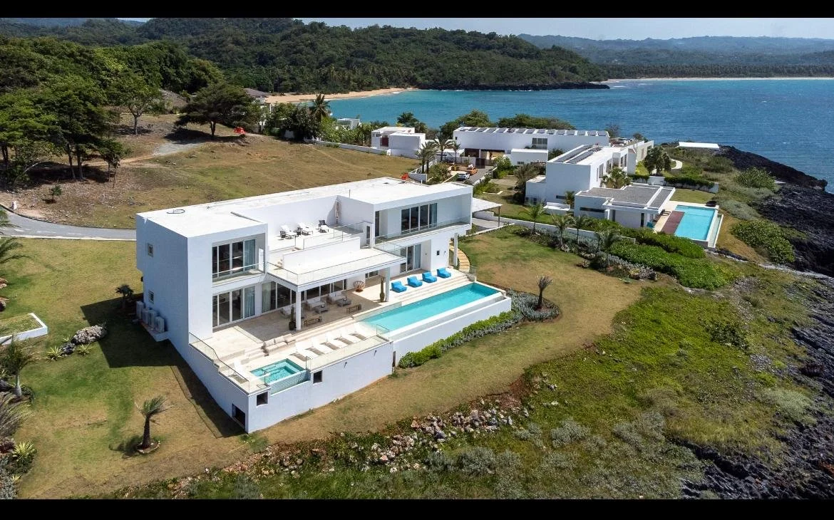 Vista aérea de un complejo residencial con casas blancas, varias piscinas y jardines, ubicado en una zona costera con playa y montañas en el fondo.