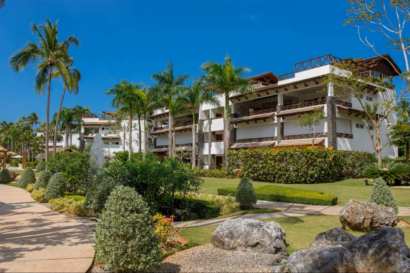 Vista de un complejo de apartamentos con jardines bien cuidados, palmeras y un cielo despejado.