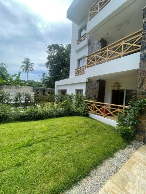 Modern three-story building with balconies and wooden railings, surrounded by a lush green lawn and trees, under a cloudy sky.