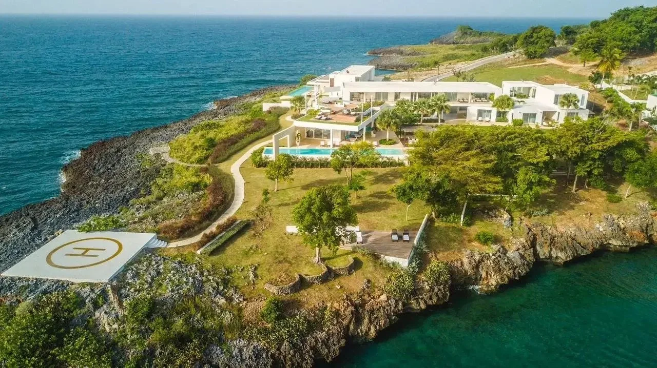 Vista aérea de un complejo de casas modernas blancas en una isla rodeada de agua, con áreas verdes y una helipuerto en la costa rocosa.