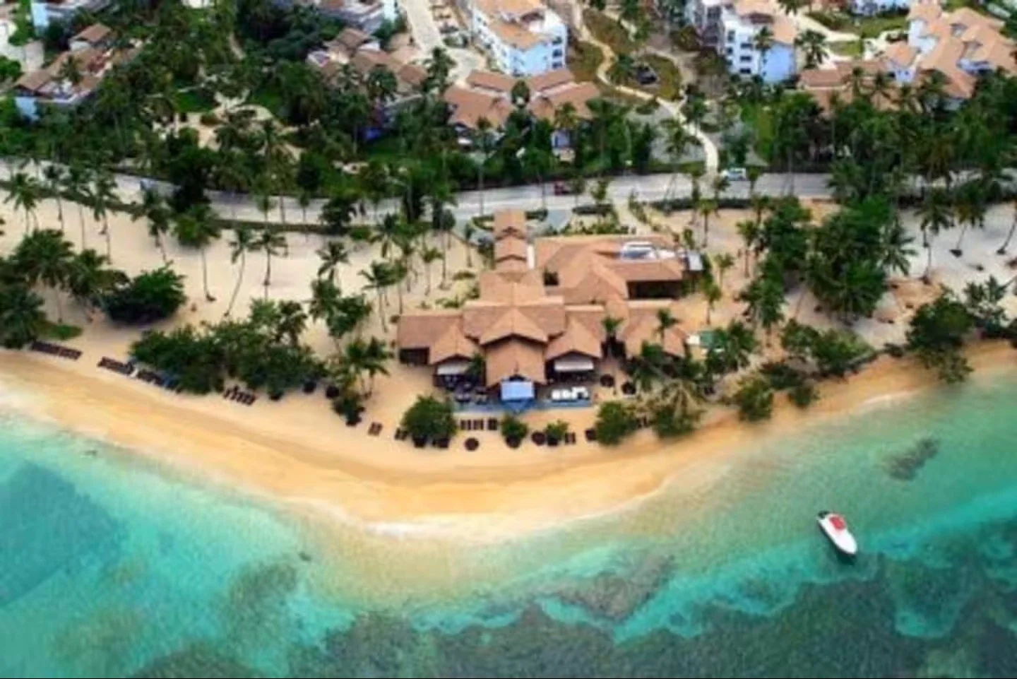 Vista aérea de un resort con casas y cabañas en la playa, rodeado de palmeras y con un bote en el agua.