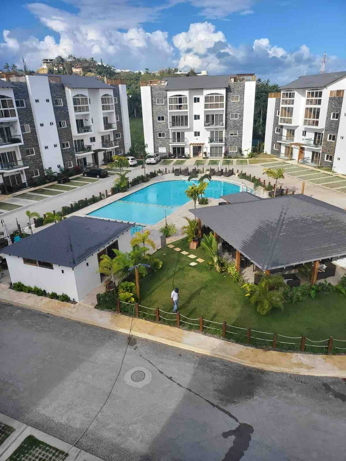 Residential complex with swimming pool, surrounded by mid-rise apartment buildings and landscaped gardens, under a blue sky with clouds.