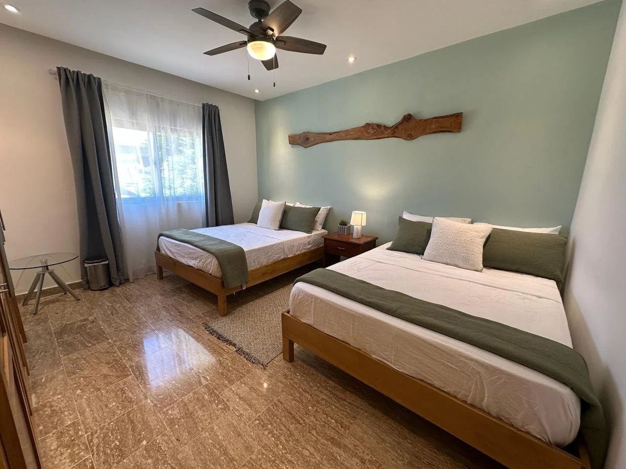 Bedroom with two double beds, green accent wall, wooden headboards, ceiling fan, window with curtains, and a nightstand with a lamp.