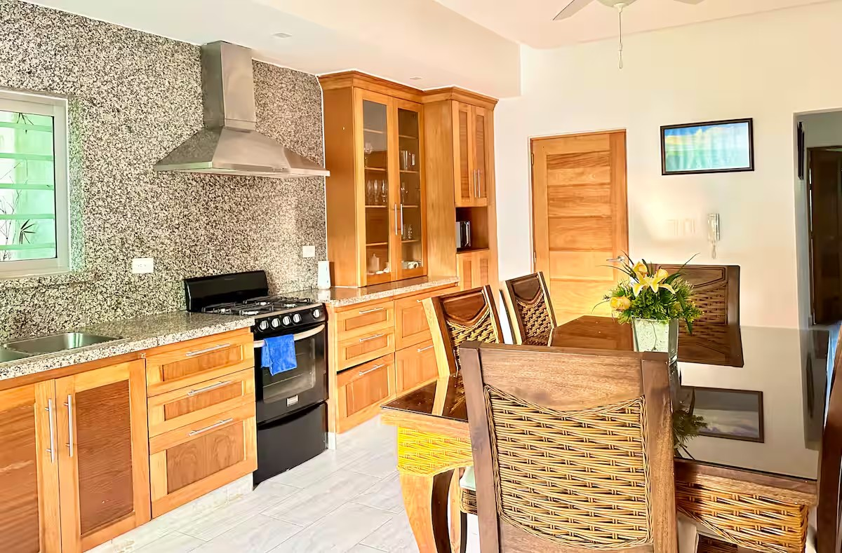 Modern kitchen with wooden cabinets, granite countertops, a stove with a stainless steel range hood, a dining table with wicker chairs, and a flower arrangement.