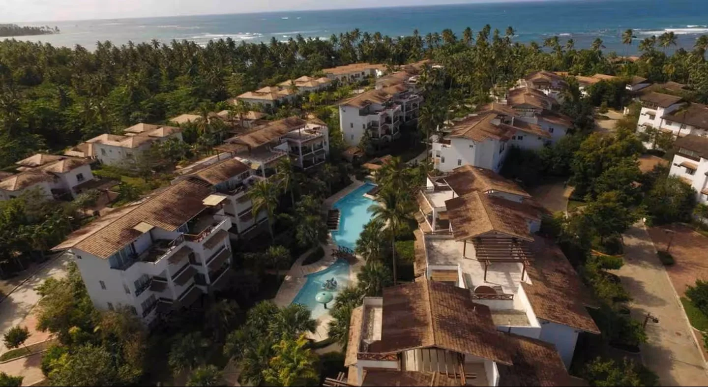Vista aérea de un complejo residencial en la playa con casas, piscinas y muchas palmeras, y el océano en el fondo.