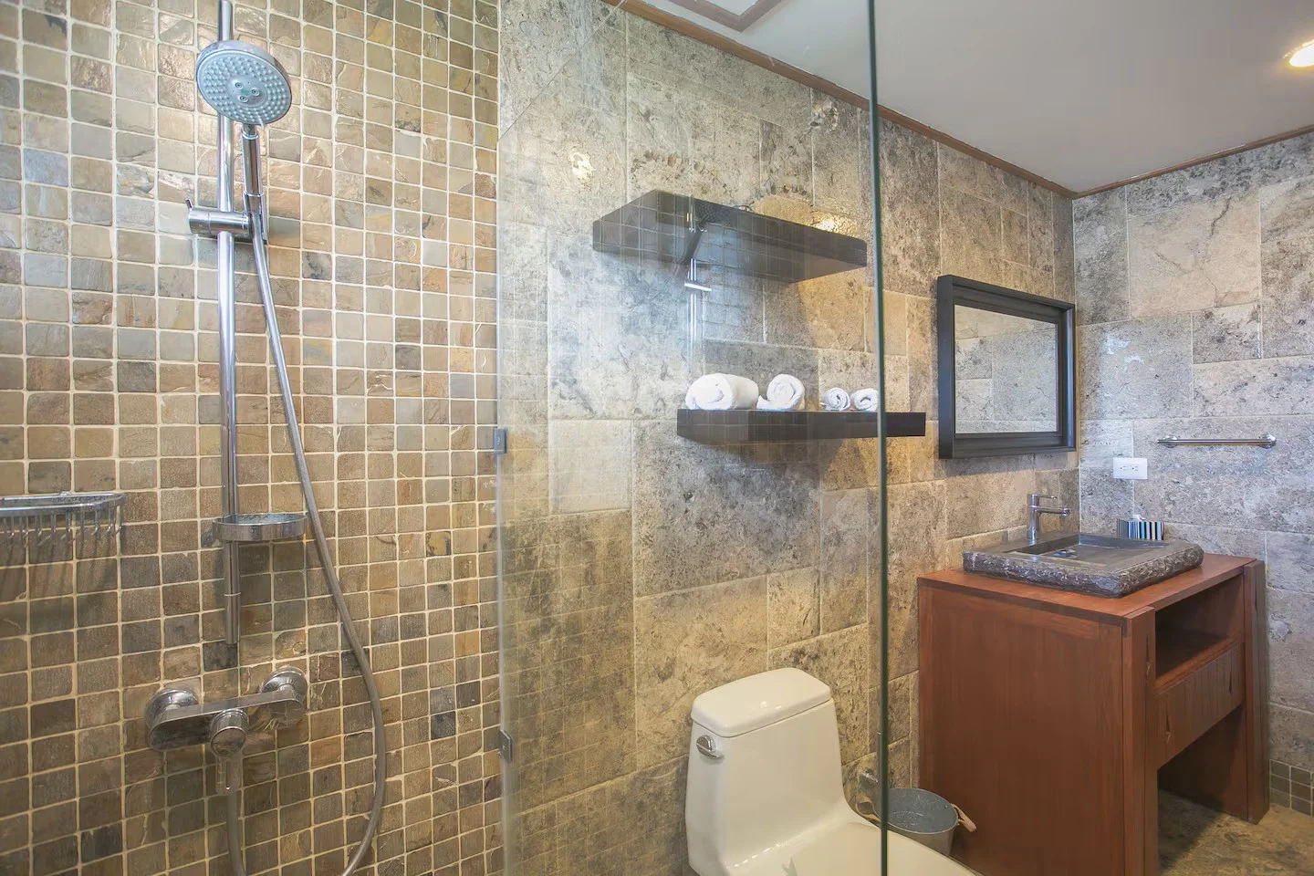 Baño de estilo rústico con paredes de piedra, ducha con azulejos pequeños y una ducha de mano, inodoro blanco y lavabo de piedra en un mueble de madera. Espejo rectangular y estantería negra con toallas, en el fondo
