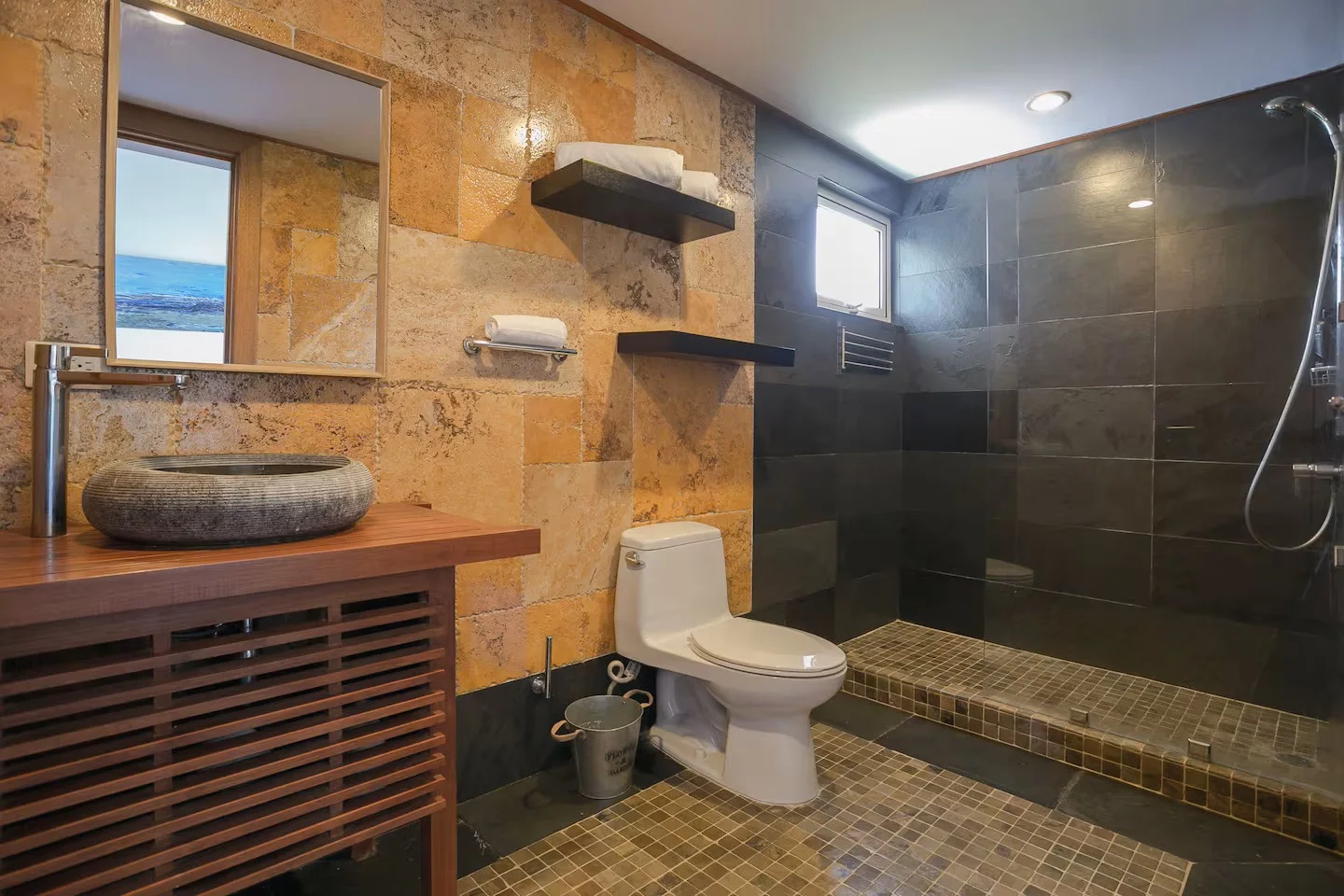 Un baño con paredes de piedra en tonos ocres y una zona de ducha con azulejos oscuros y suelo de mosaico, un lavabo de piedra sobre una superficie de madera y un espejo rectangular.
