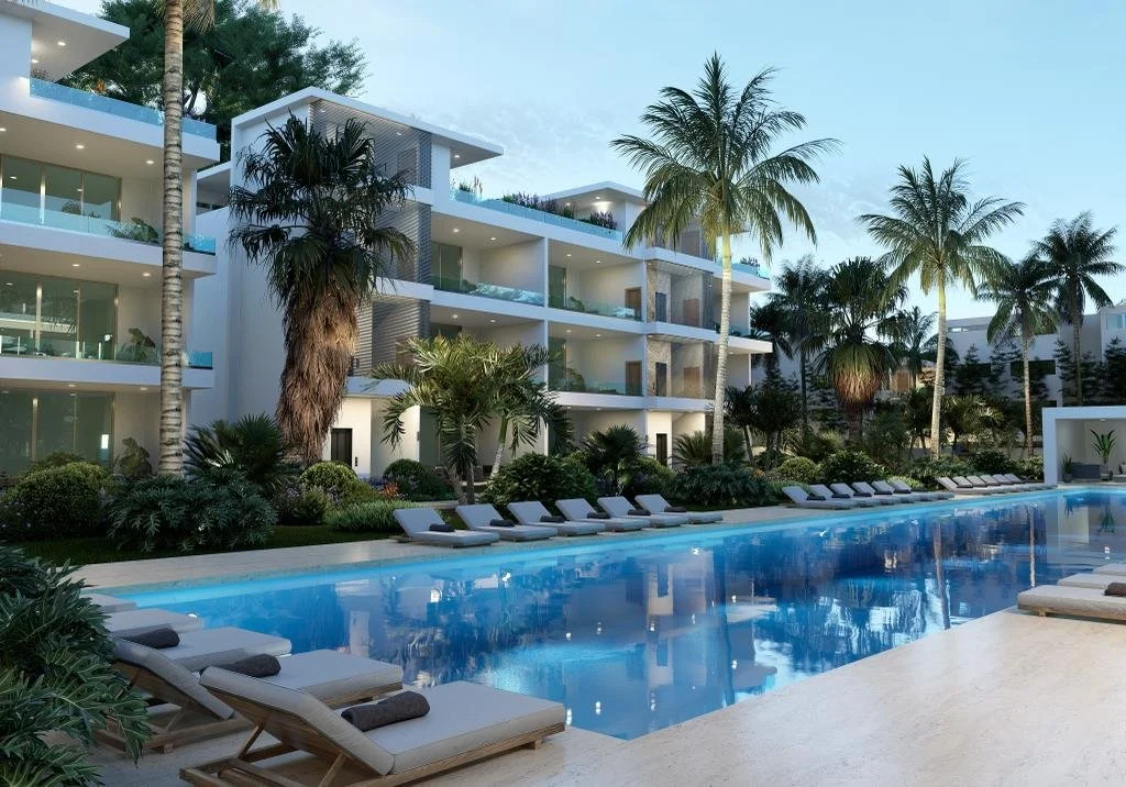 Complejo de apartamentos moderno con piscina, rodeado de palmeras y tumbonas, bajo un cielo despejado.