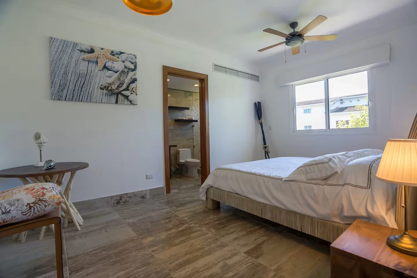 Habitación con cama, mesita de noche con lámpara, ventana grande, cuadros en la pared, vista del baño y una escalera en una esquina.