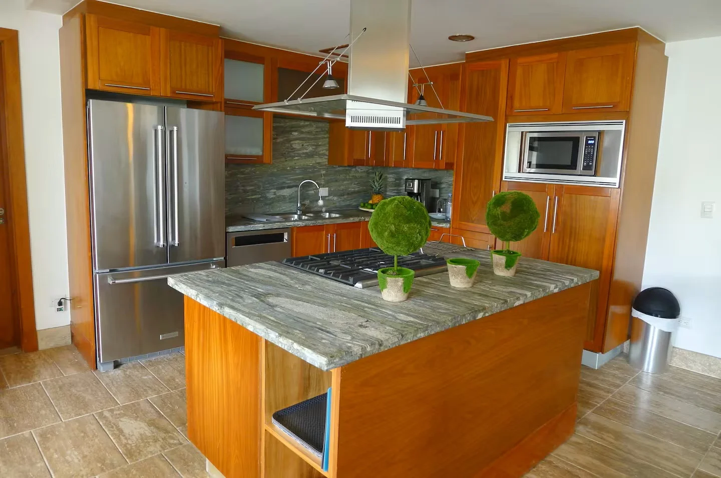 Cocina moderna con gabinetes de madera, encimera de piedra, electrodomésticos de acero inoxidable, y decoraciones verdes en la isla central.