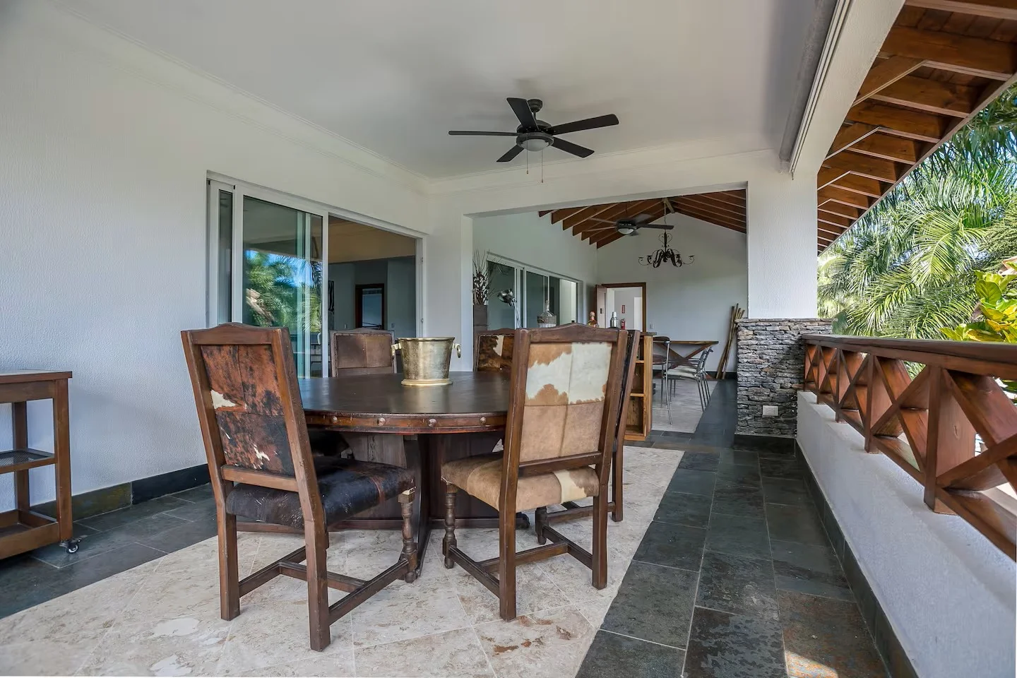 Amplio balcón con mesa de comedor de madera, sillas de diferentes estilos, techo de madera con ventiladores, paredes blancas, piso de piedra, y vista a la vegetación tropical.