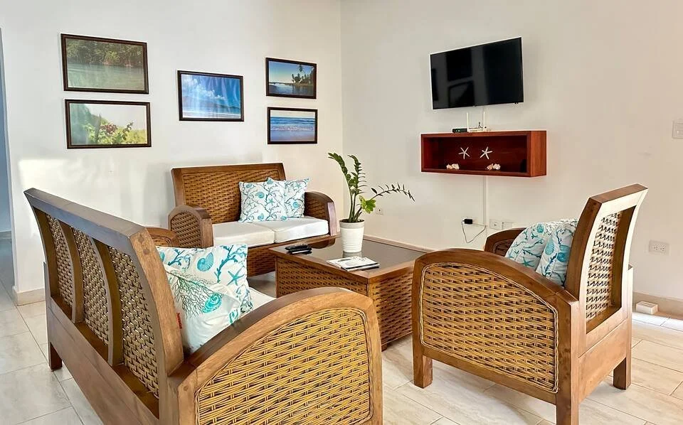 Cozy living room with wooden furniture, white cushions, floral pillows, wall-mounted TV, nature-themed wall art, and indoor plant.