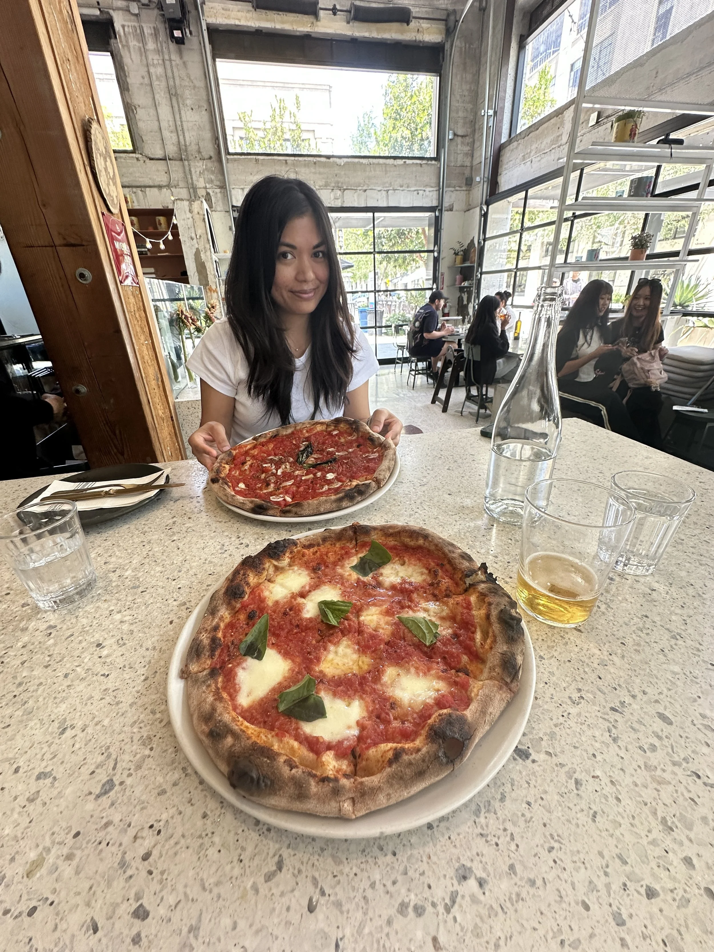 A woman holding a pizza in a restaurant with large windows and other diners in the background. Another pizza and a half-full glass of beer are on the table.