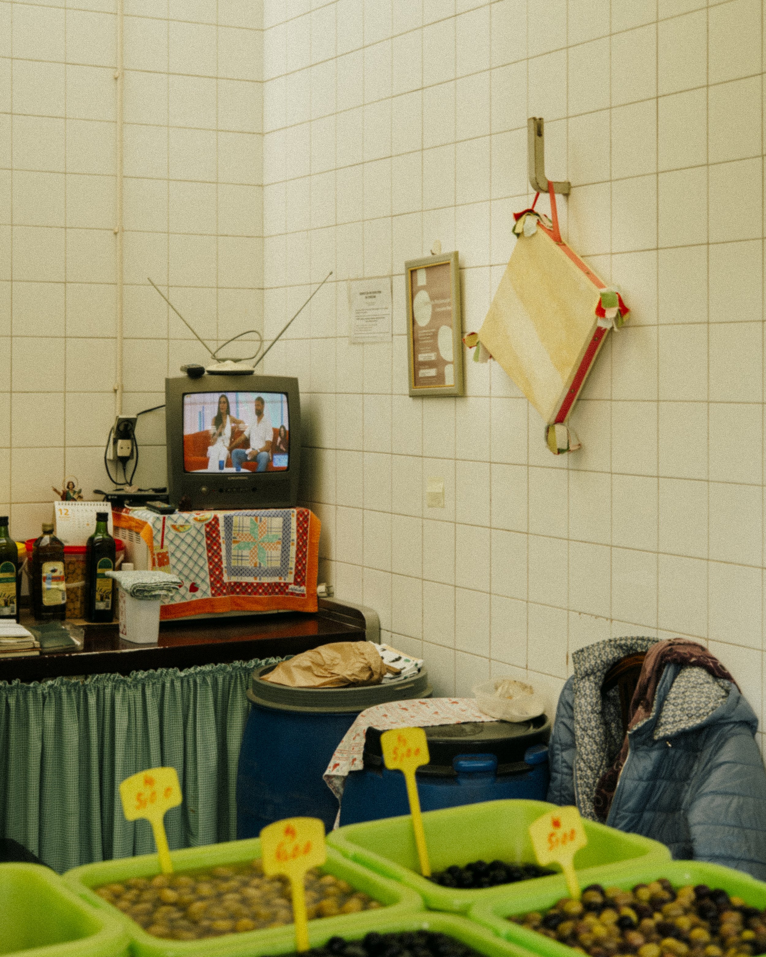 Interior de uma loja com televisores, garrafas, roupas e caixas de frutas com preços, parede de azulejos e objetos pendurados.