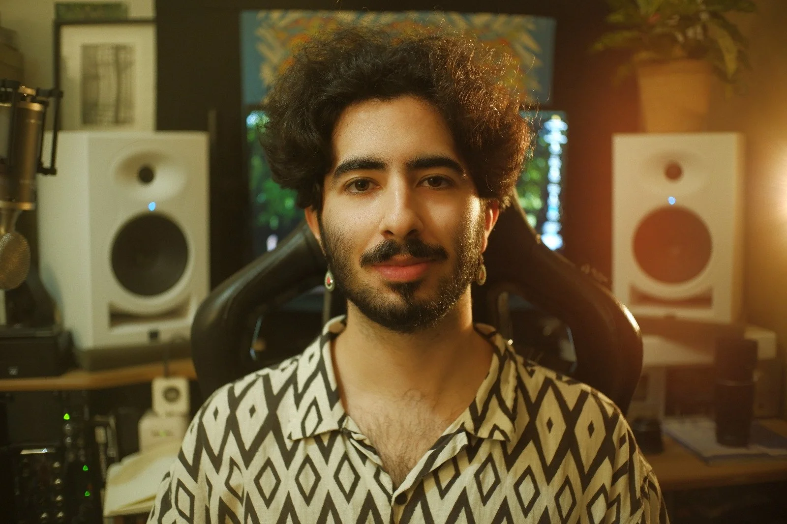 Homem com cabelo afro, barba, usando uma camisa com padrão geométrico, sentado em um estúdio de gravação, com monitores de áudio e equipamento de estúdio ao fundo, iluminação quente.