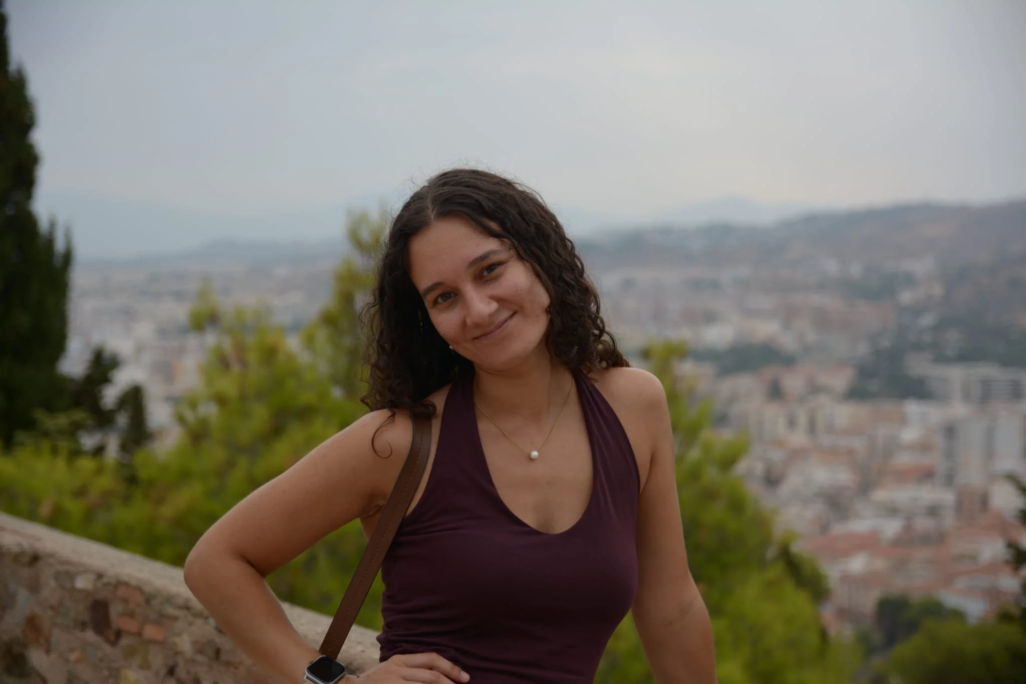 Mulher sorrindo, usando top roxo e colar de pérola, com cabelo cacheado, ao ar livre com cidade ao fundo.