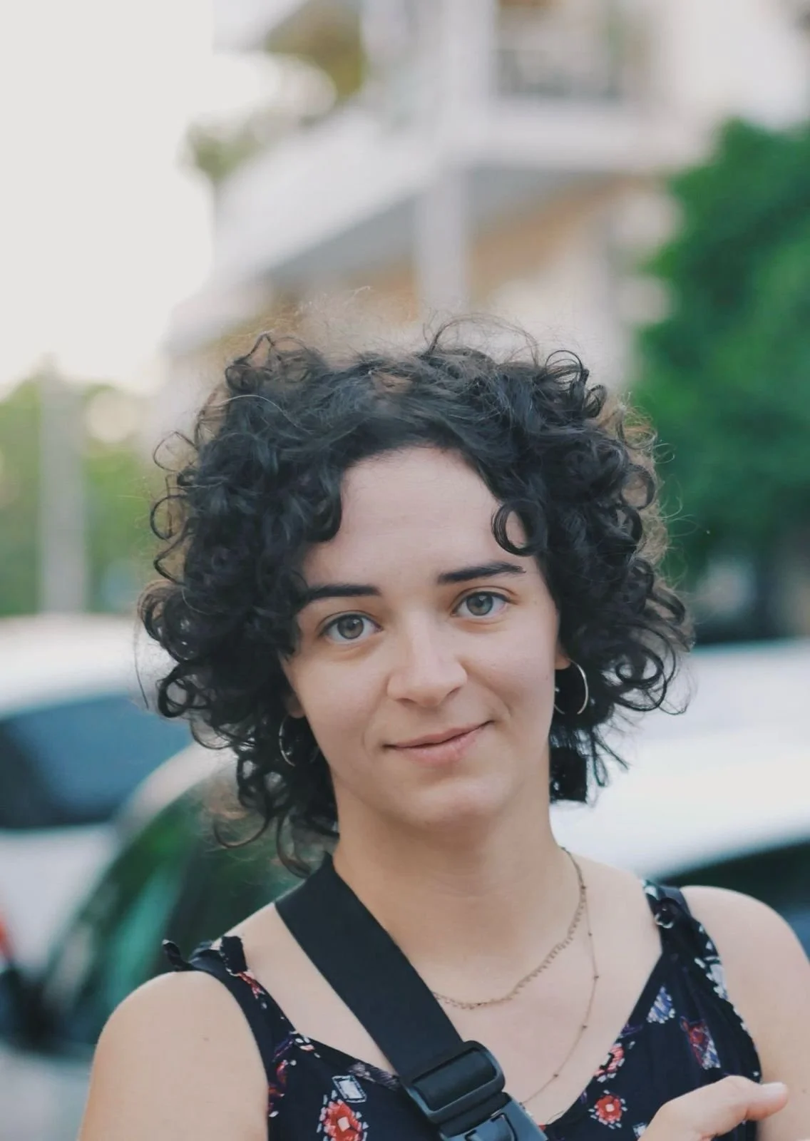 Jovem mulher com cabelo cacheado, pele clara, usando uma camiseta preta com estampa floral e uma mochila, sorrindo na rua com carros ao fundo.