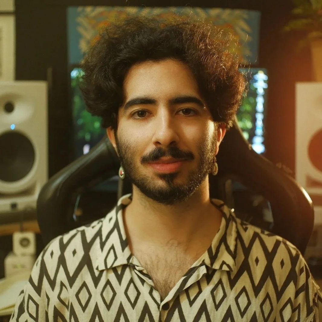 Homem com cabelo e barba escuros e cacheados, usando camisa com padrão geométrico, sentado em sala de estúdio com equipamentos de áudio e iluminação.