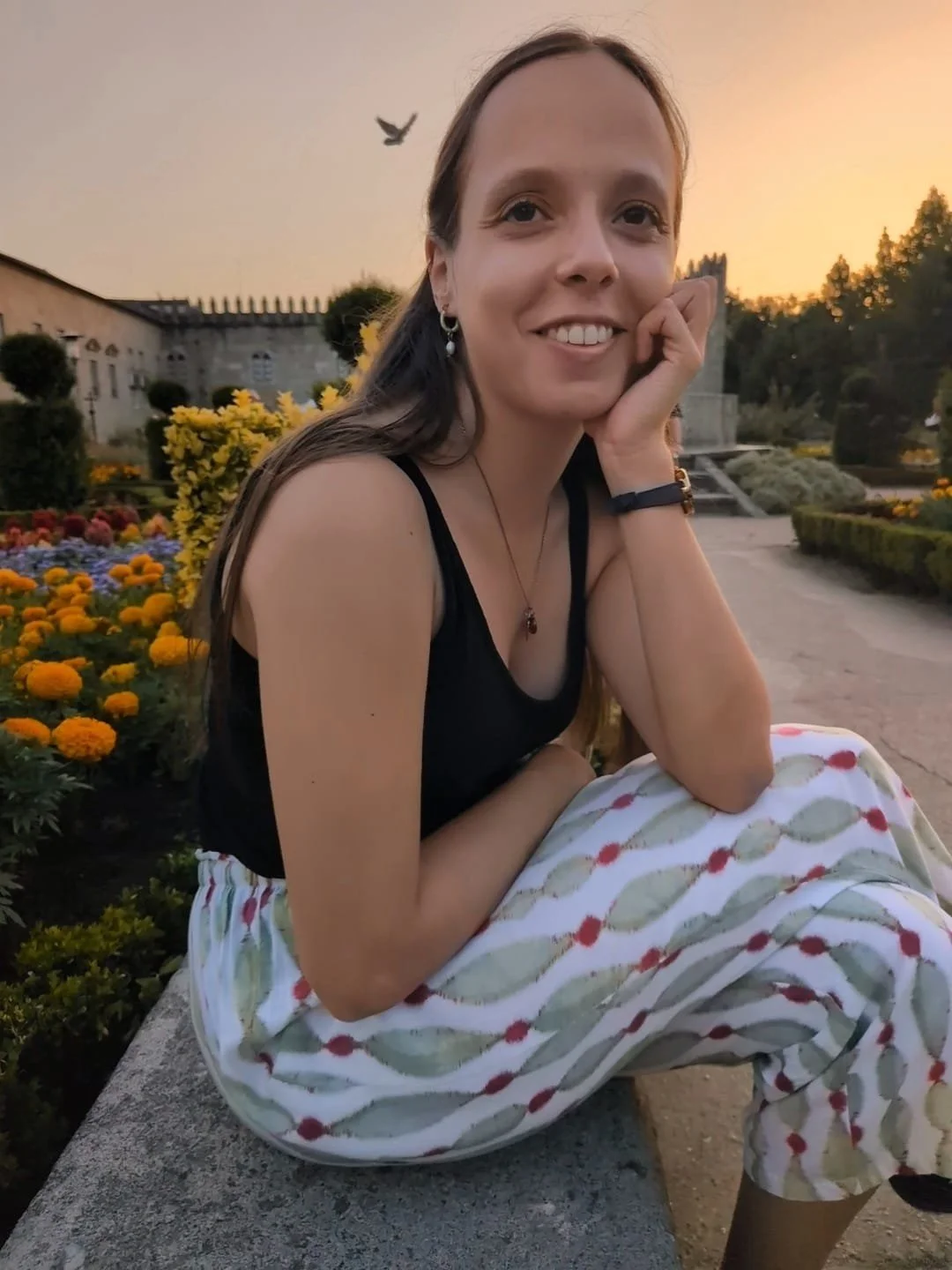 Jovem sorridente com roupas casuais, sentada em banco de pedra em jardim com flores e árvores ao entardecer, com pássaro voando ao fundo.