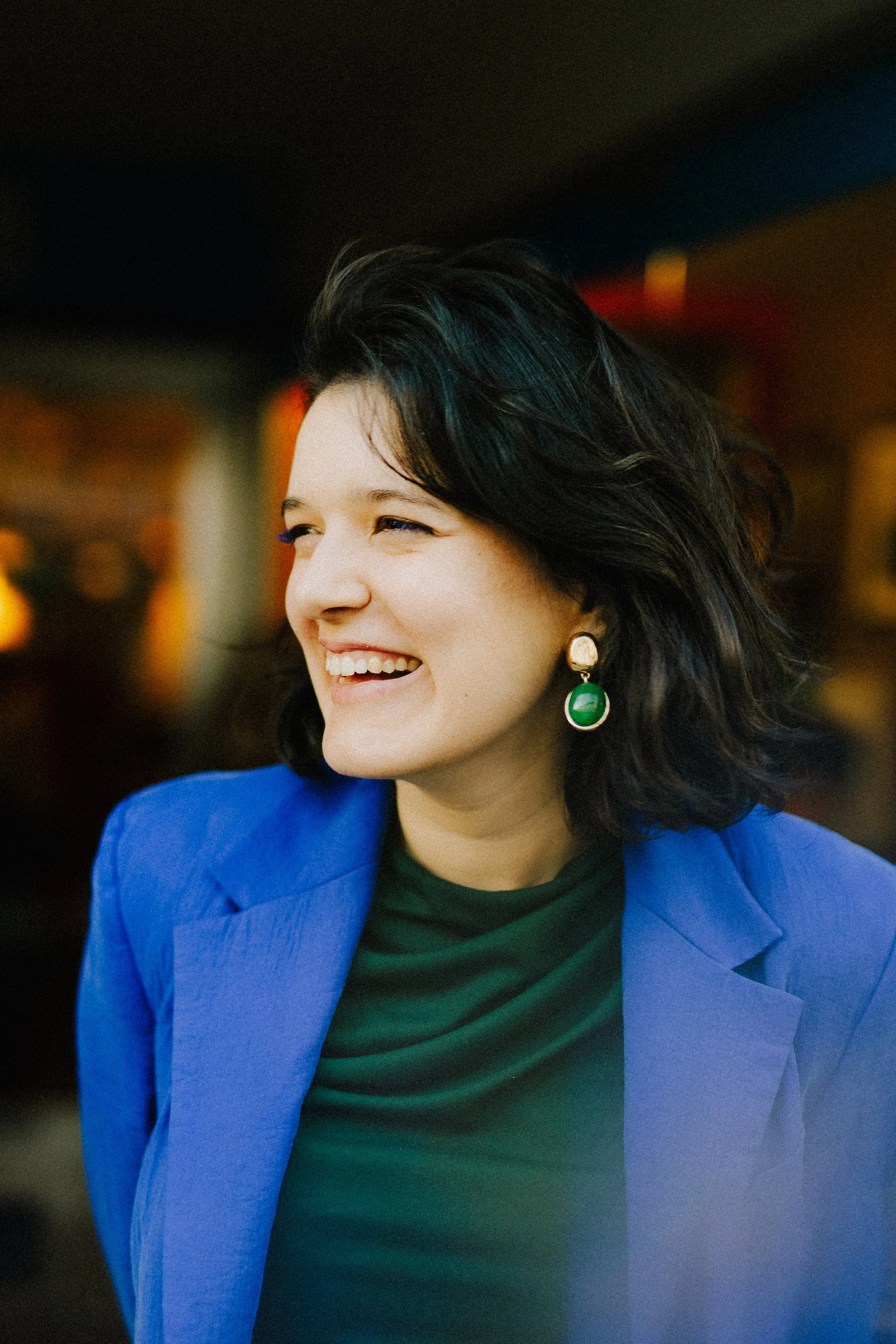 Mulher sorrindo vestida com blazer azul e blusa verde, usando brincos grandes verdes, com cabelo preto e ondulado, ambiente com iluminação quente ao fundo.