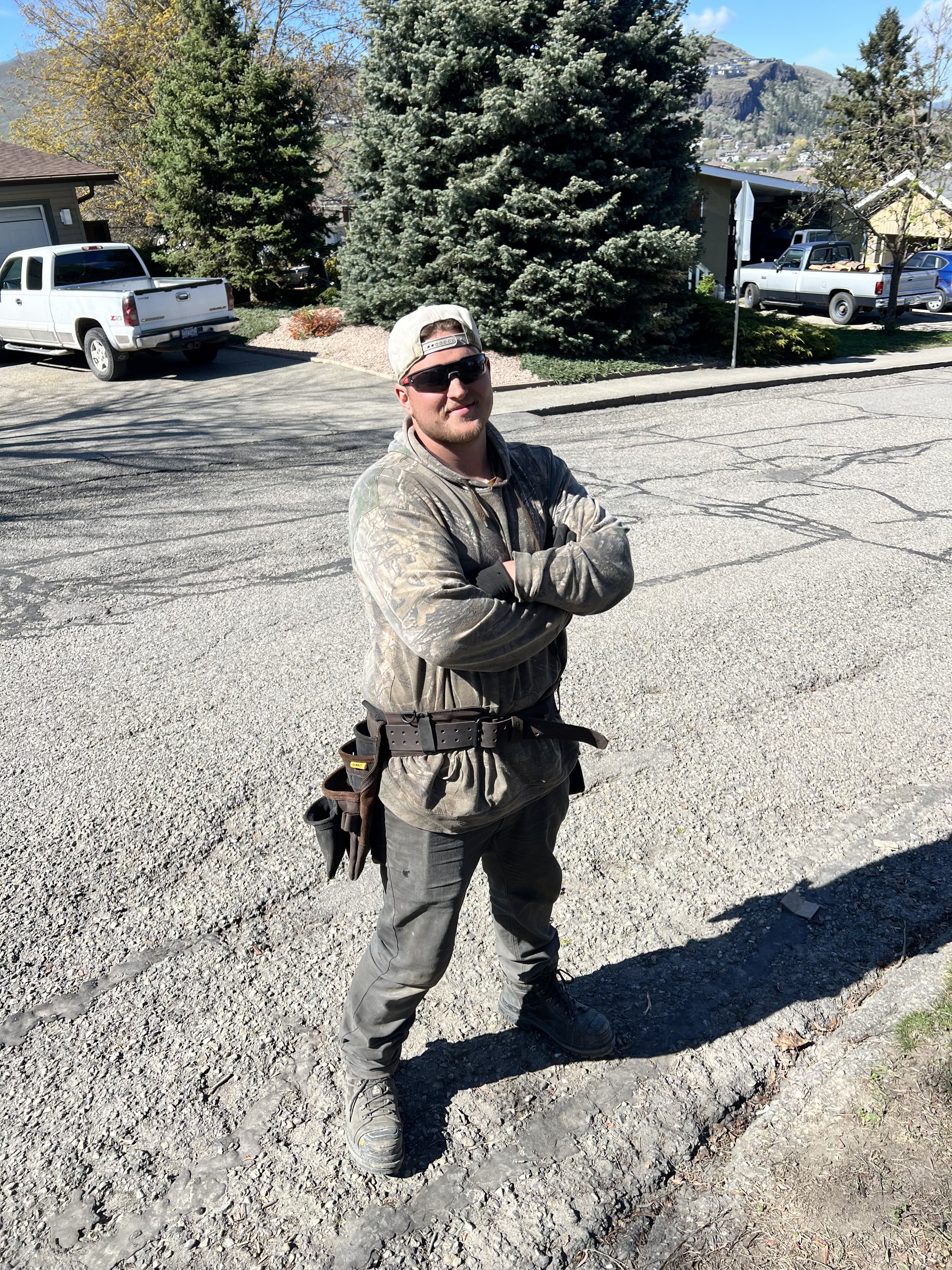 A man in outdoor gear, wearing sunglasses and a baseball cap, standing on a gravel street with arms crossed. He has a tool belt and kneepads on, with residential houses, parked trucks, and pine trees behind him. Mountain in the background.
