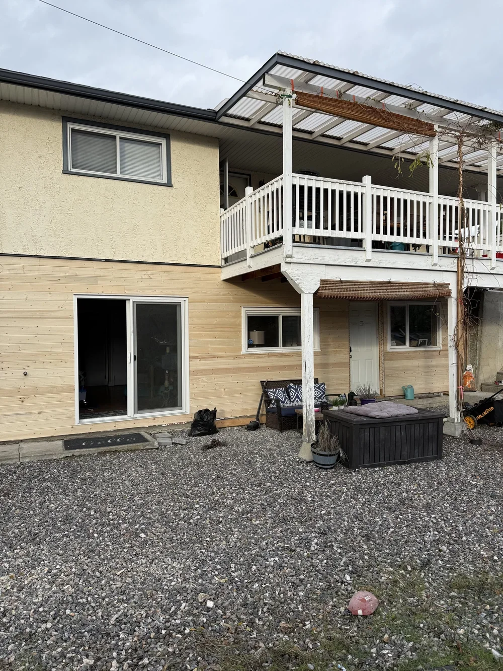 Back view of a two-story house with a gravel yard, featuring a partially covered balcony, outdoor seating, and a storage chest.