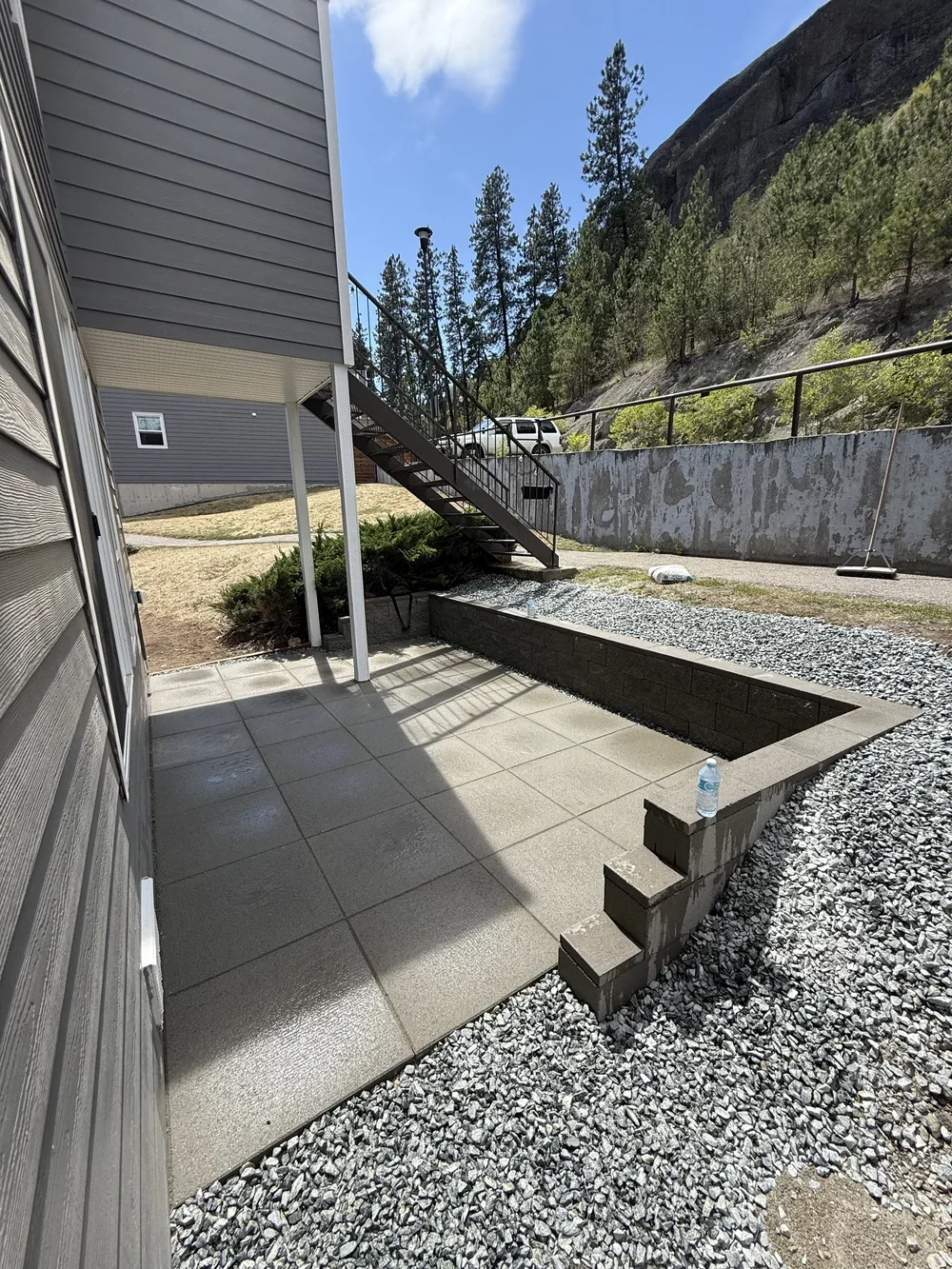 A small outdoor patio area with concrete tiles beside a house, with stairs leading up to a second story. The patio is bordered by a low wall with gravel outside it, and a plastic water bottle sits on one of the steps. In the background, there is a gravel pathway, a slope with pines and trees, and a concrete retaining wall with a black metal railing at the top.