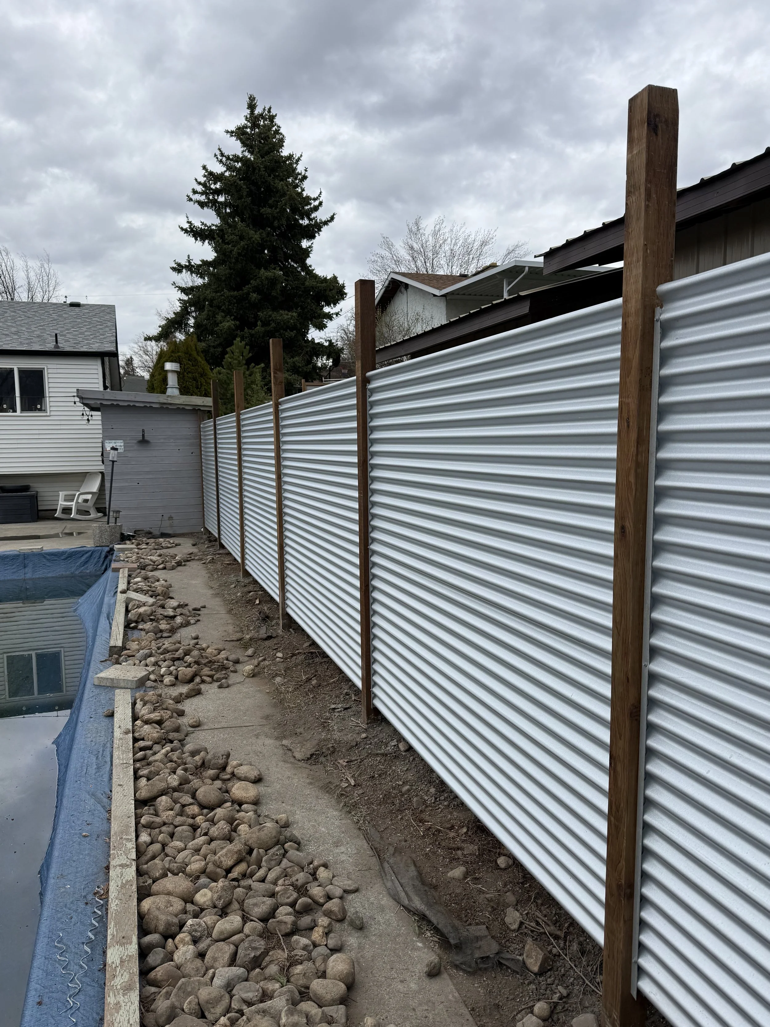 A new corrugated metal fence supported by wooden posts along a backyard, with a swimming pool on the left and residential houses in the background under cloudy sky.