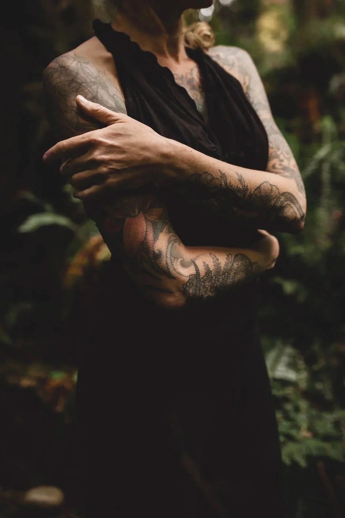 A woman with twisted arms crossing her chest, displaying tattoos on her arms and chest, wearing a dark sleeveless top, outdoors in a natural setting.