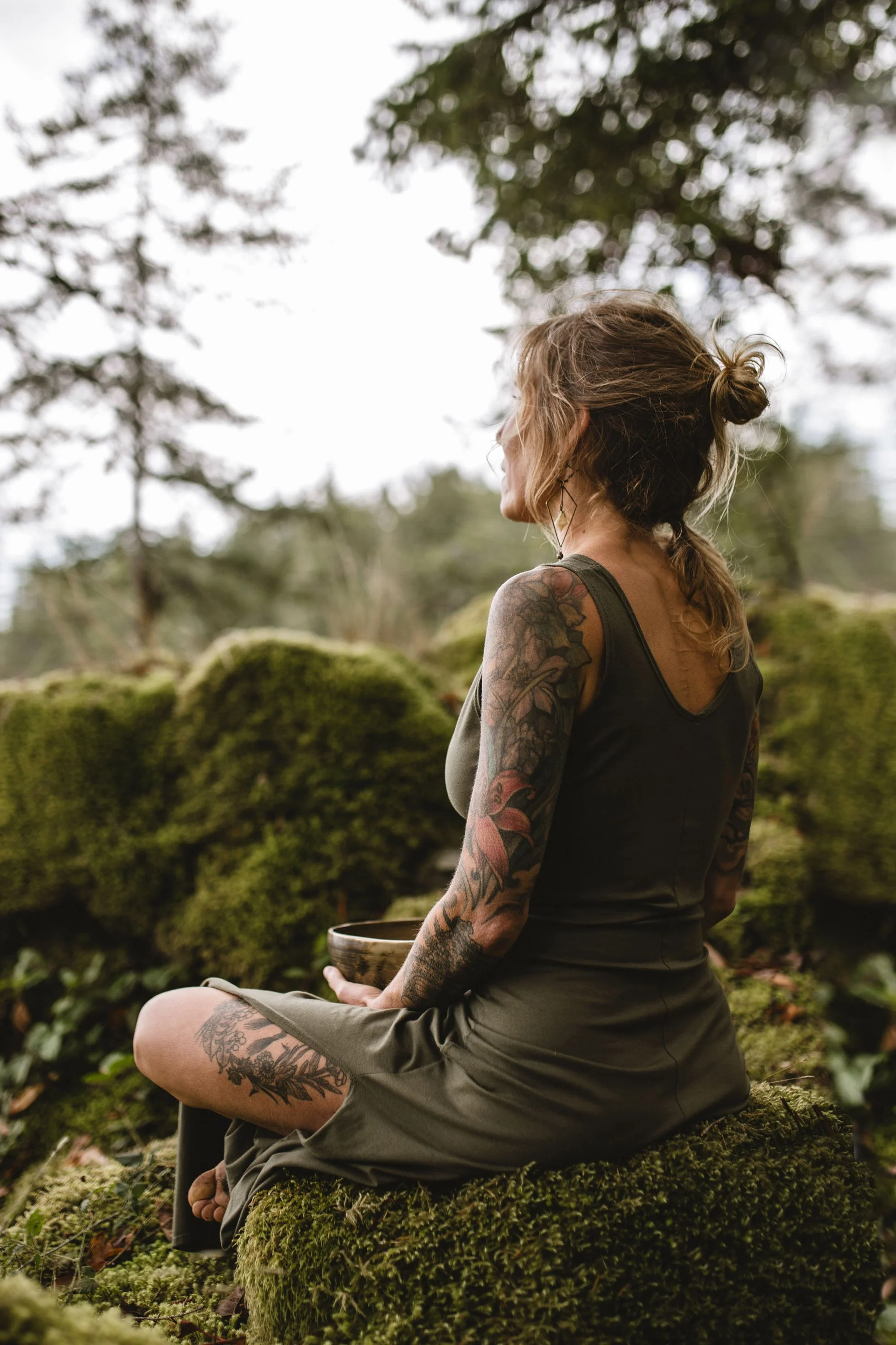 Nataliesits on a moss-covered rock in a forest, holding a small bowl, her back turned toward the camera.