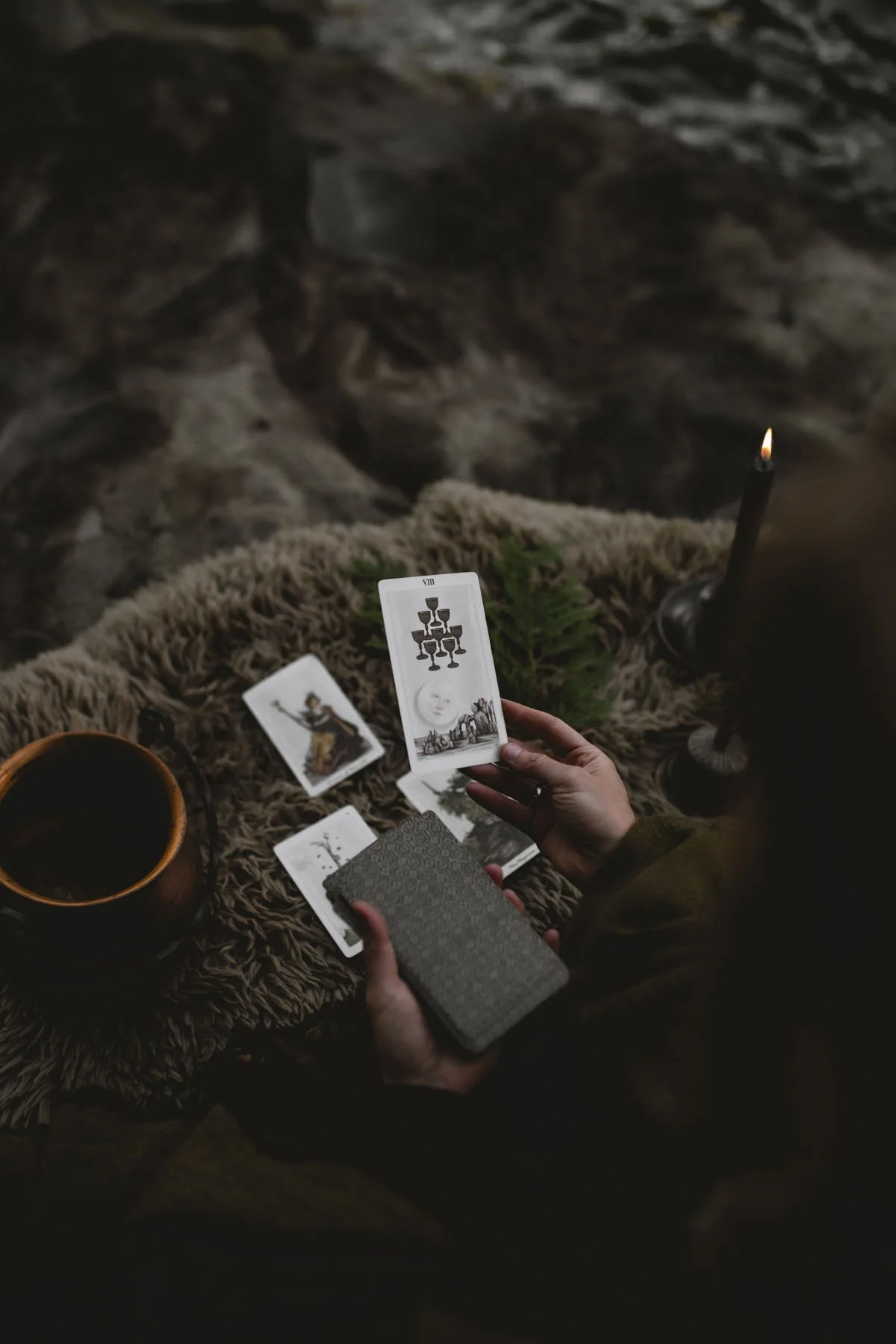 A person holding tarot cards over a furry blanket with a candle, a cup of coffee, and a small green plant on a rustic wooden surface.